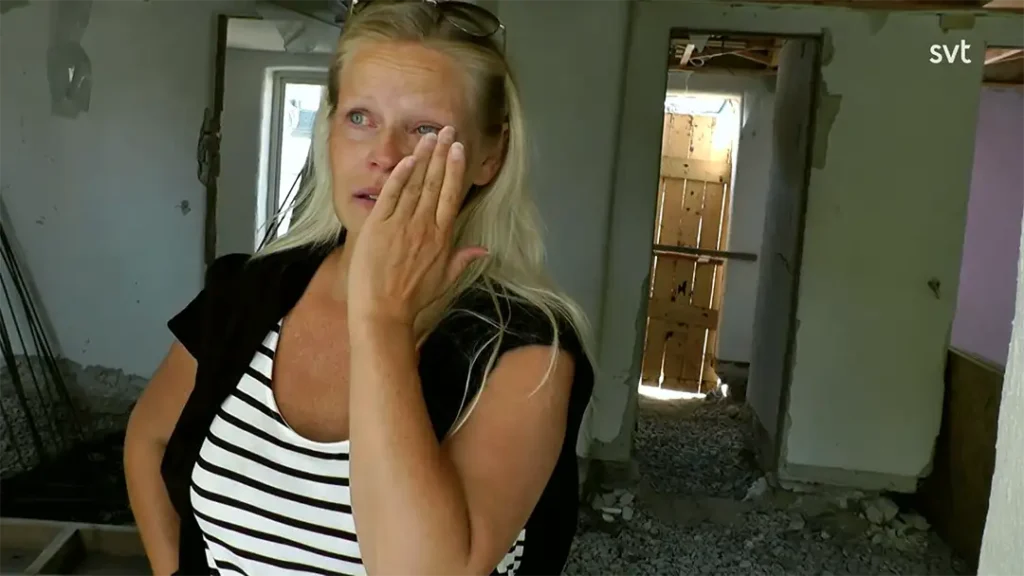 Stiligt - En kvinna med långt blont hår torkar ögat när hon står i ett skadat, oavslutat rum med skräp på golvet.