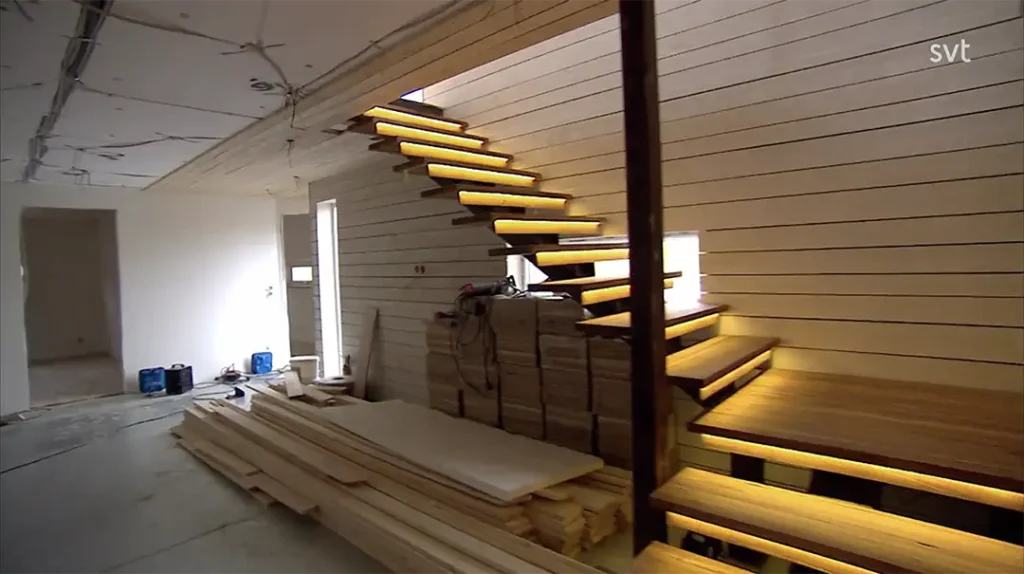 Stiligt - En modern interiör under uppbyggnad i Västra Hamnen - Husdrömmar stil har en svävande trätrappa med inbyggda lampor; träplankor och byggmaterial ligger utspridda på golvet.