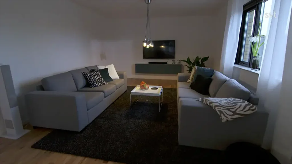 Stiligt - Ett modernt vardagsrum inspirerat av Furulund - Husdrömmar, med två grå soffor, en svart matta, ett litet soffbord, vägghängd TV och ett stort fönster med skira vita gardiner.