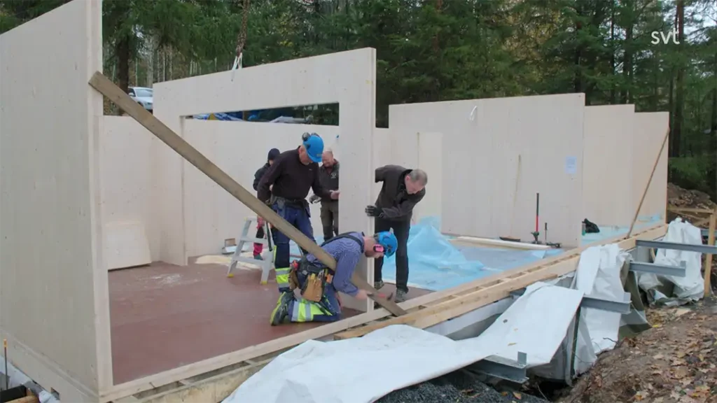 Stiligt - I Värmland monterar fyra personer träväggskivor till en Husdrömmarsbyggnad under uppförande i ett skogsområde, med verktyg och material utspridda på platsen.