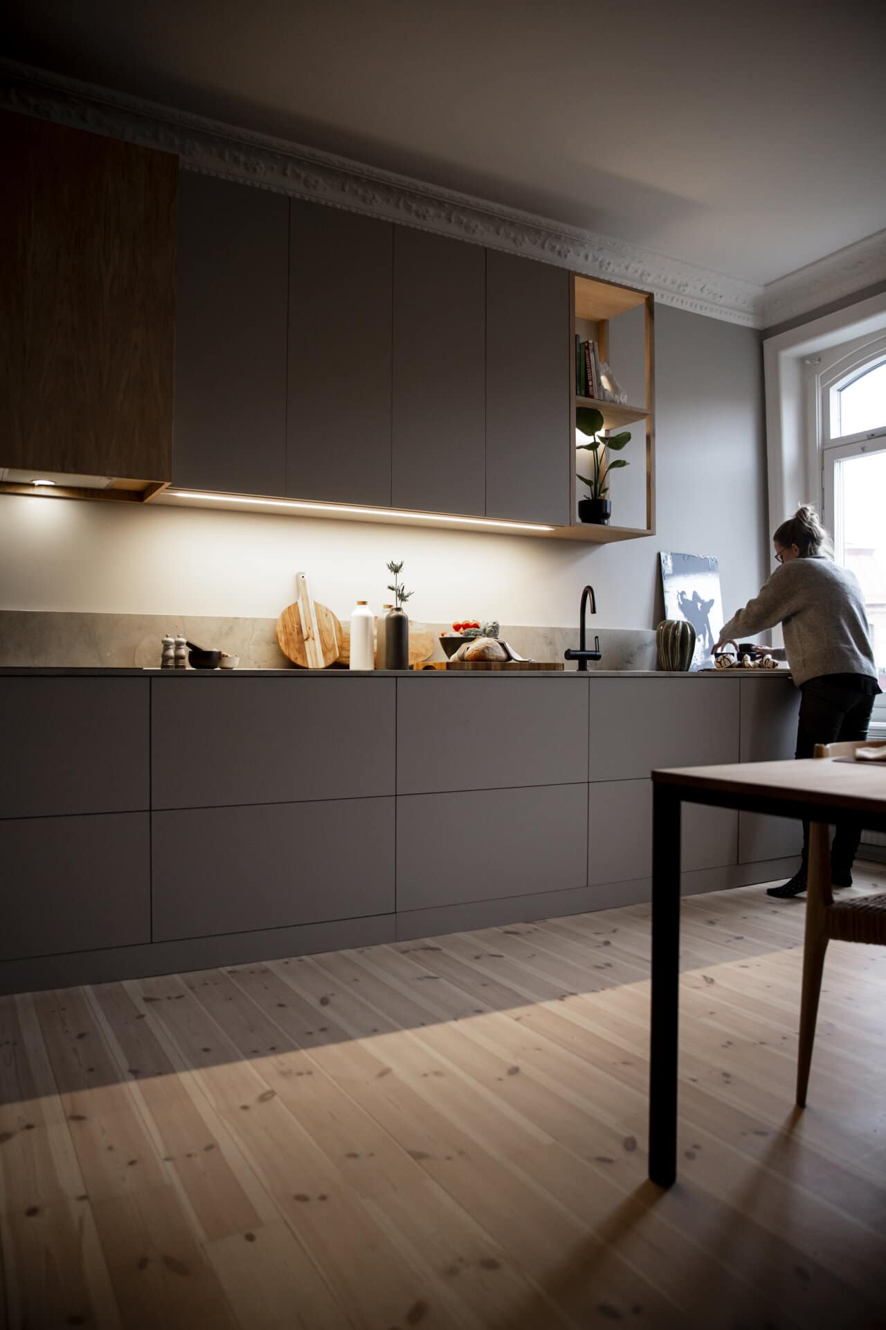 Stiligt - En person står vid en diskbänk i ett modernt, platsbyggt kök med gråa skåp, belysning under skåpet och trädetaljer.