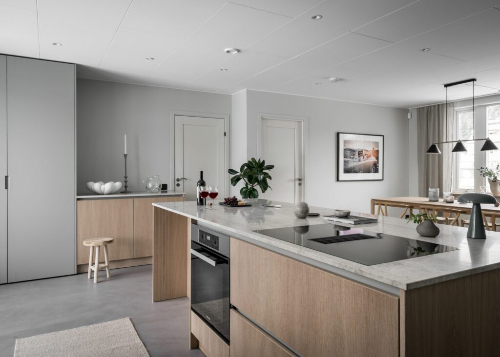 Stiligt - Modernt kök med ljusa träskåp, bänkskivor i marmor, inbyggda vitvaror, en central ö och en intilliggande matplats med stora fönster och minimalistisk inredning.