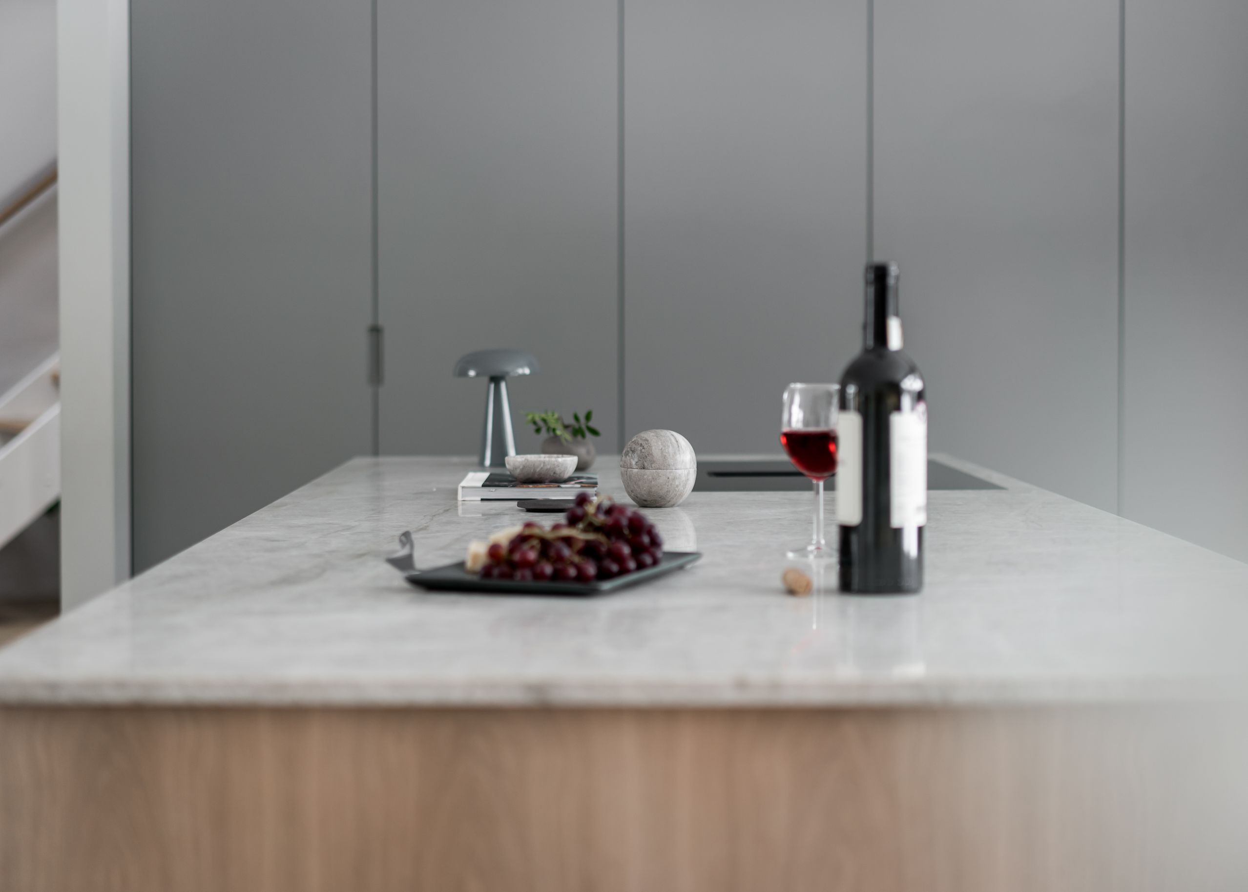 Stiligt - En modern köksö med en bänkskiva i stiligt marmor visar upp en flaska rött vin, ett glas, vindruvor på en tallrik, en liten lampa och dekorativa föremål.