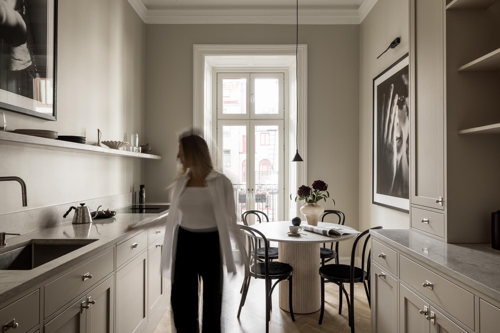 Stiligt - En person står i en kök i shakerstil med ljusa skåp, ett runt matbord med stolar och stora fönster som släpper in dagsljus.