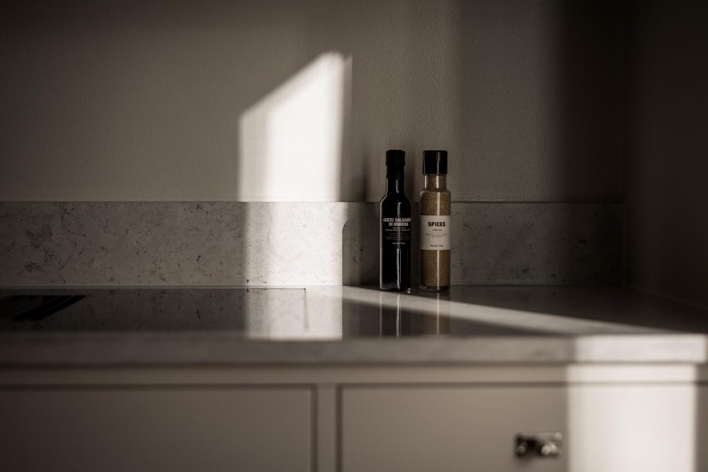 Stiligt - Två flaskor, en mörk och en ljus, sitter på en bänkskiva i marmor i en kök i shakerstil, när solljuset strömmar in och kastar mjuka skuggor på vägg och yta.