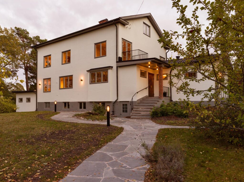 Stiligt - Ett tvåvånings krämfärgat hus med flera fönster, en utomhustrappa som leder till entrén, en stengång mitt bland en gräsmatta och träd och en stilren träkök med pocketdörrar inuti.