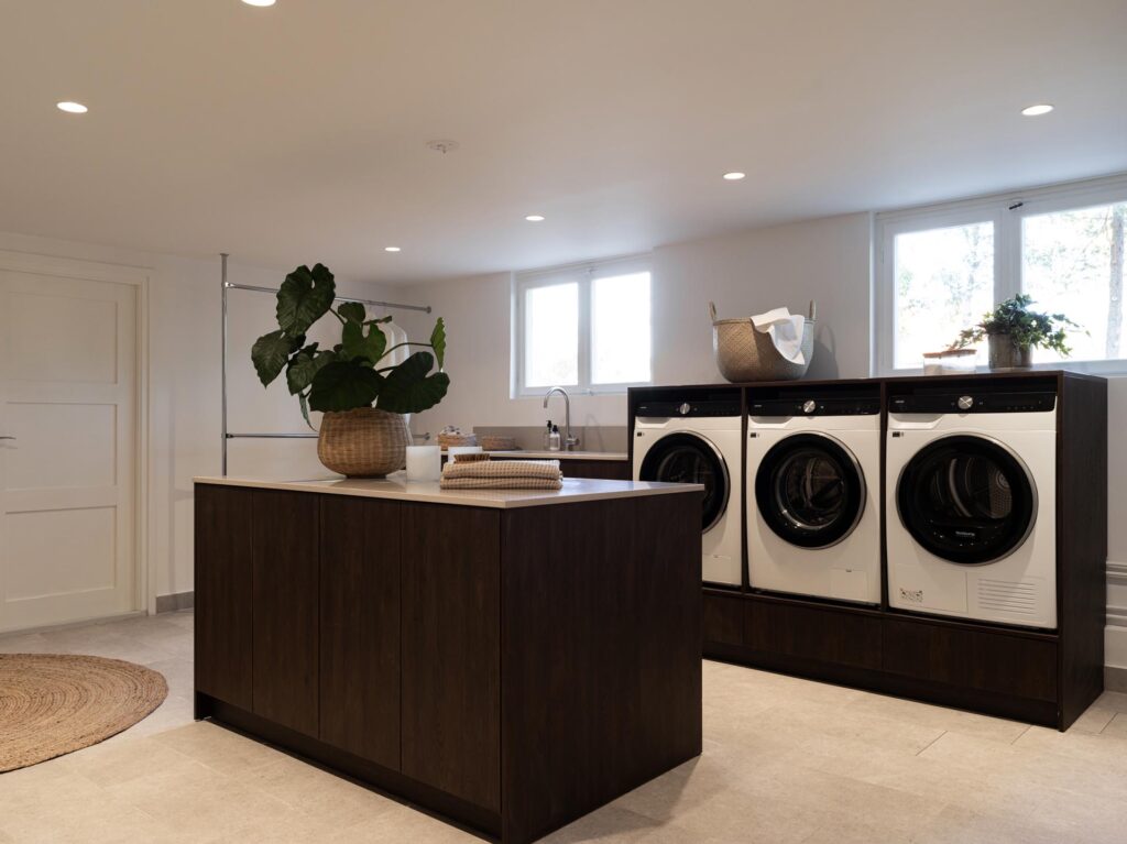 Stiligt - Modern tvättstuga med tre frontmatade maskiner, en mörk träö med prydligt vikta handdukar, en korg och en planta, under starkt takbelysning intill stora fönster och en träkök med pocketdörrar.