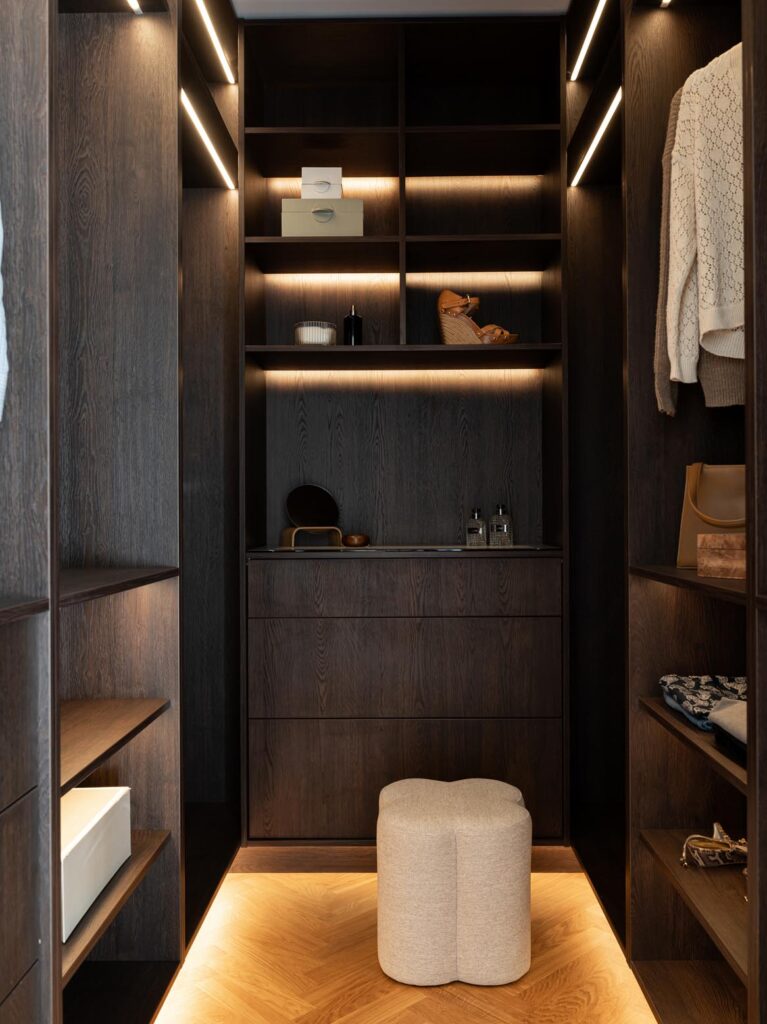 Stiligt - En modern klädkammare med hyllor, lådor och belysning i mörkt trä – utformad som en träkök med pocketdörrar – med snyggt arrangerade kläder, lådor, accessoarer och en beige ottoman på ett trägolv.