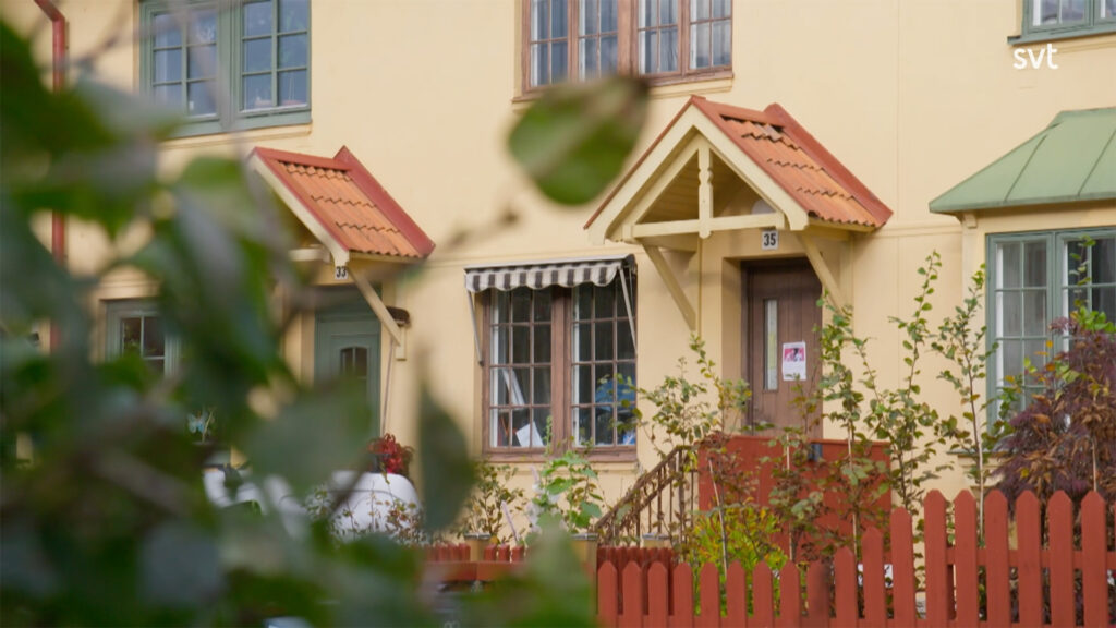 Stiligt - Charmigt gult hus med rött staket, liten veranda, och fönstermarkis i Gamla Enskede - Husdrömmar stil, delvis dolt av grönt bladverk. Hus nummer 35 syns nära dörren.