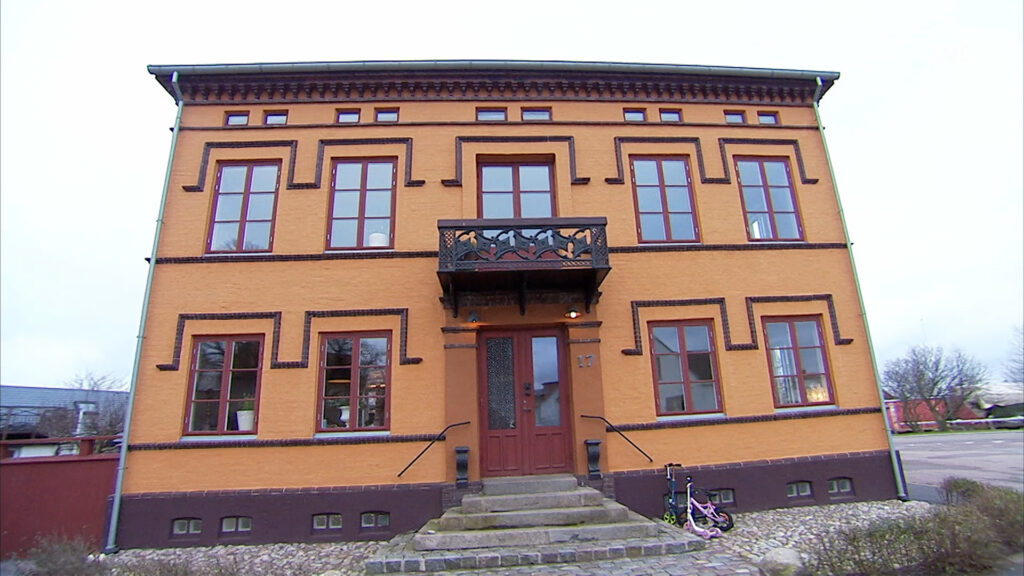 Stiligt - Tvåvåningshus i orange och brunt tegel med symmetriska fönster, central trädörr, balkong ovanför entrén, stentrappa och en rosa barncykel vid trappan.