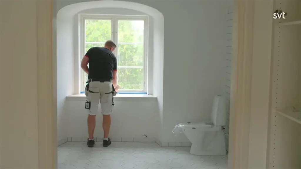 Stiligt - En person står nära ett fönster med verktyg i handen i ett vitkaklat badrum, med toalett och ser ut att vara under renovering, som en del av projektet Södra Björstorp - Husdrömmar.
