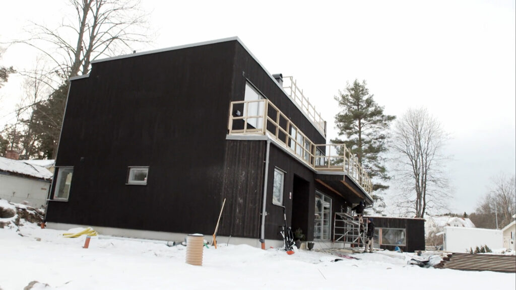 Stiligt - Ett modernt svart trähus med stora fönster under uppbyggnad, omgivet av snö och utspridda byggmaterial på marken.