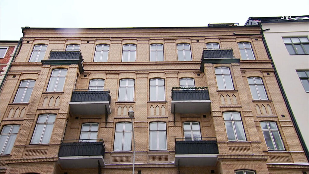 Stiligt - Ett hyreshus i fyra våningar med stora fönster och flera svarta metallbalkonger, sett från gatuplan.