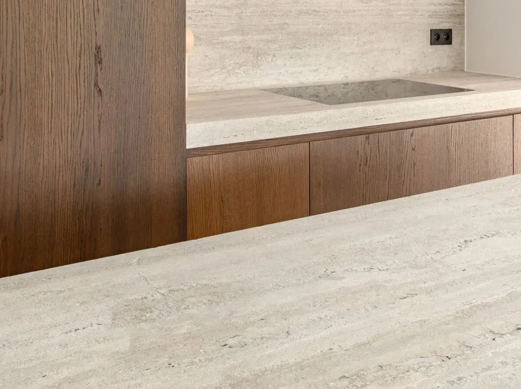 Stiligt - Minimalistiskt kök med ljus stenbänkskiva, integrerad spishäll, träskåp och beige texturerad stänkskydd. Två eluttag syns på väggen.