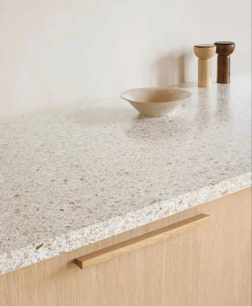 Stiligt - En bänkskiva i terrazzo med en beige skål och två träbägare ovanpå, placerad ovanför ett ljust träskåp med ett horisontellt handtag.